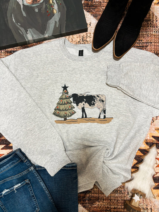 Light grey crewneck with a longhorn steer and Christmas tree graphic, styled flat lay with jeans and cow painting – western holiday sweatshirt.