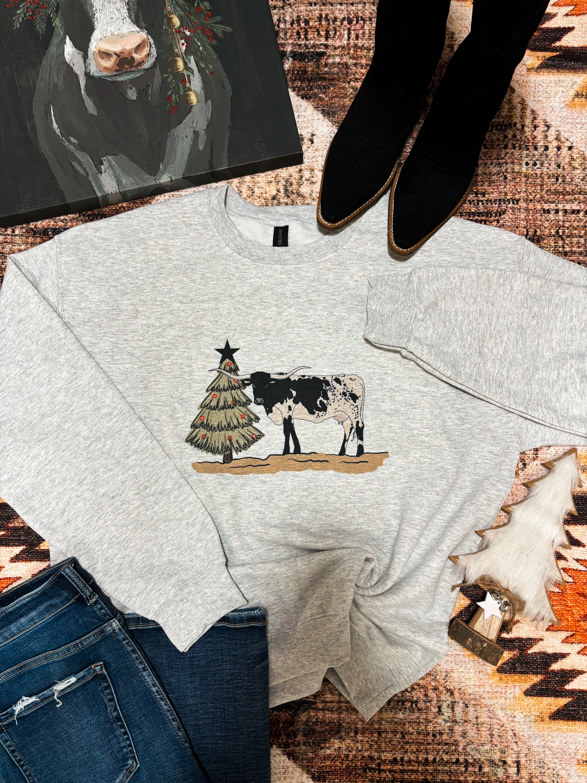 Longhorn cow and rustic Christmas tree printed on grey sweatshirt, styled with denim and woodsy decor – festive country crewneck.