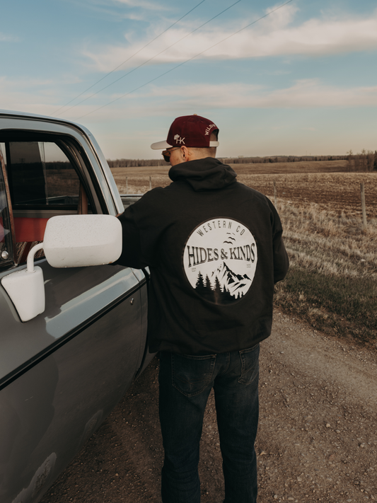 Men's black Western Co hoodie by Hides & Kinds featuring a large mountain back graphic, photographed beside a vintage truck on a country road.