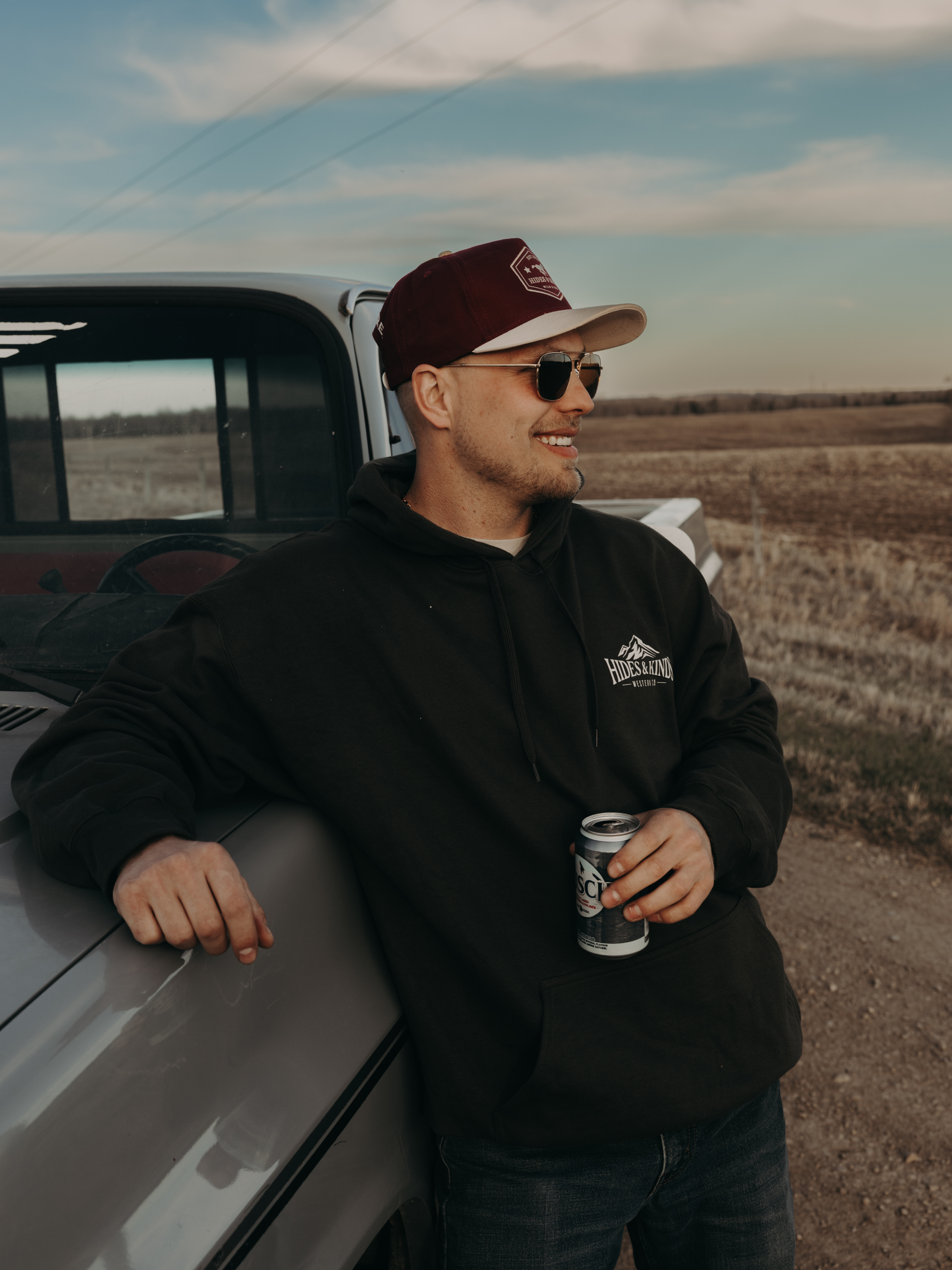 Man wearing the black Hides & Kinds Western Co hoodie with small chest logo, leaning against a vintage truck in a rural setting.