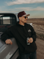 Man wearing the black Hides & Kinds Western Co hoodie with small chest logo, leaning against a vintage truck in a rural setting.