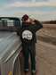 Man wearing the Hides & Kinds High Country Hoodie in Smoke with the back graphic design, standing beside a vintage pickup truck on a country road.