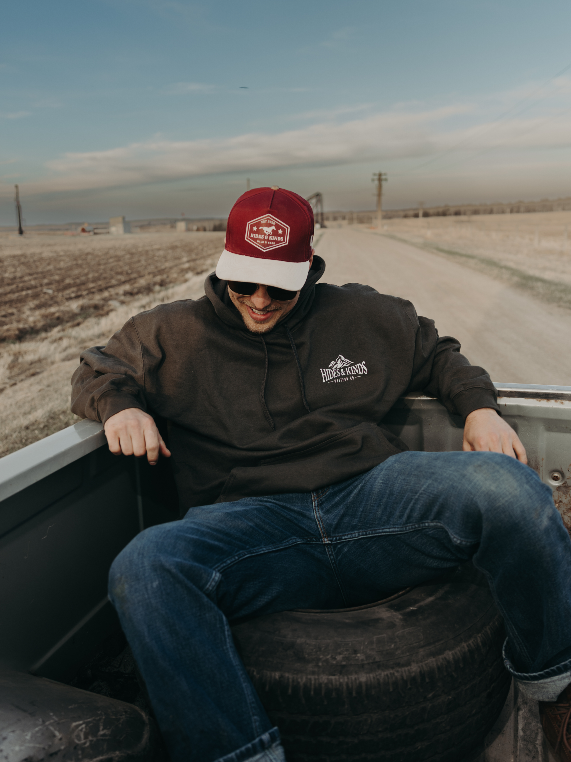 Man wearing the Hides & Kinds High Country Hoodie in Smoke while sitting in the bed of a vintage pickup truck on a country road.