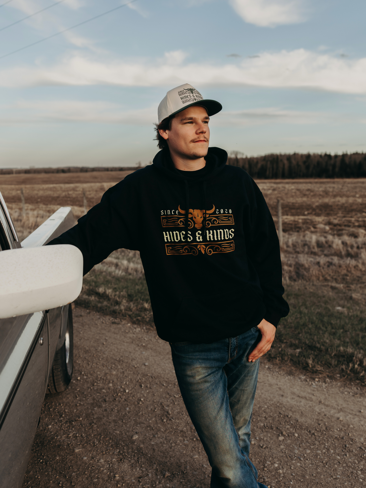 Man wearing the Hides & Kinds Men's Ranchline Hoodie in black with the front western logo design, standing beside a vintage pickup truck on a country road.