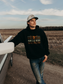Man wearing the Hides & Kinds Men's Ranchline Hoodie in black with the front western logo design, standing beside a vintage pickup truck on a country road.