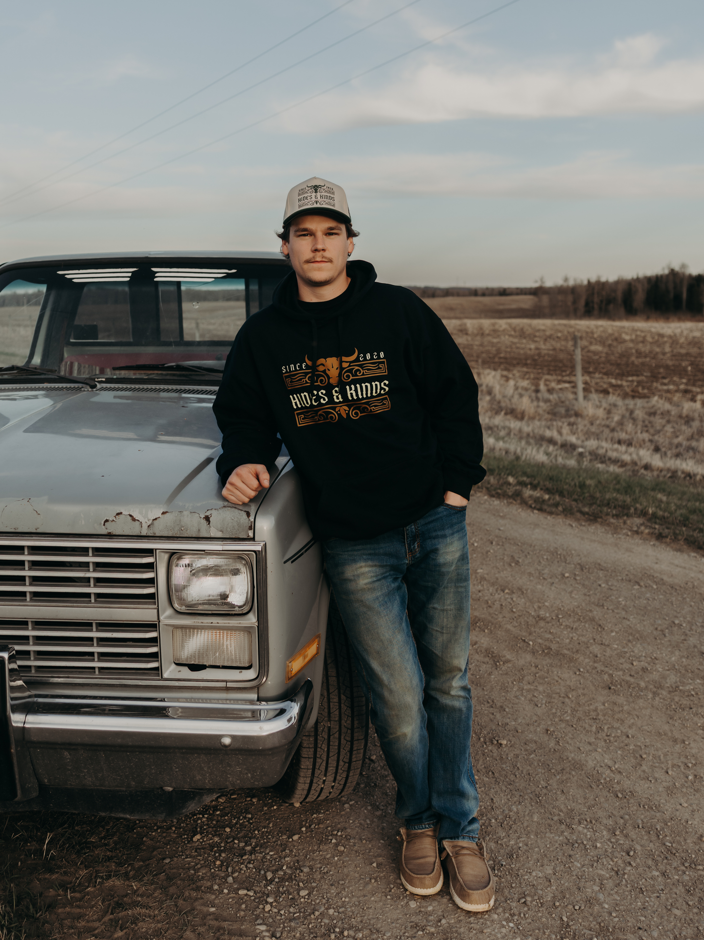 Full-body photo of a man wearing the Hides & Kinds Men's Ranchline Hoodie in black with the front logo design, leaning against a vintage pickup truck in the countryside.
