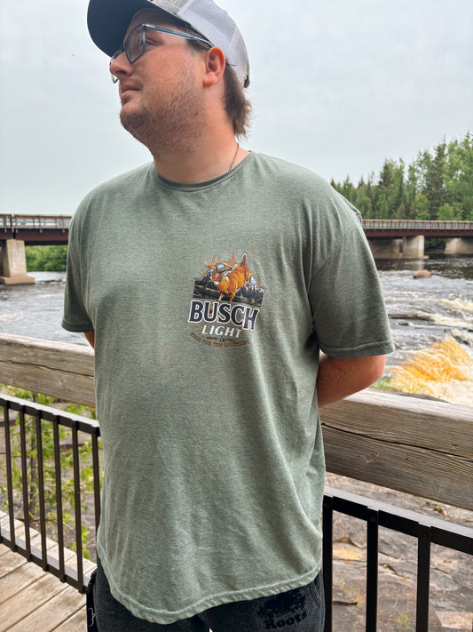Man wearing the Mountain Buck Busch Tee with deer and mountain design on a sage green t-shirt near a river