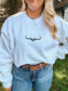 Lifestyle shot of the Original Logo Crew in a backyard setting, styled with jeans and silver jewelry.