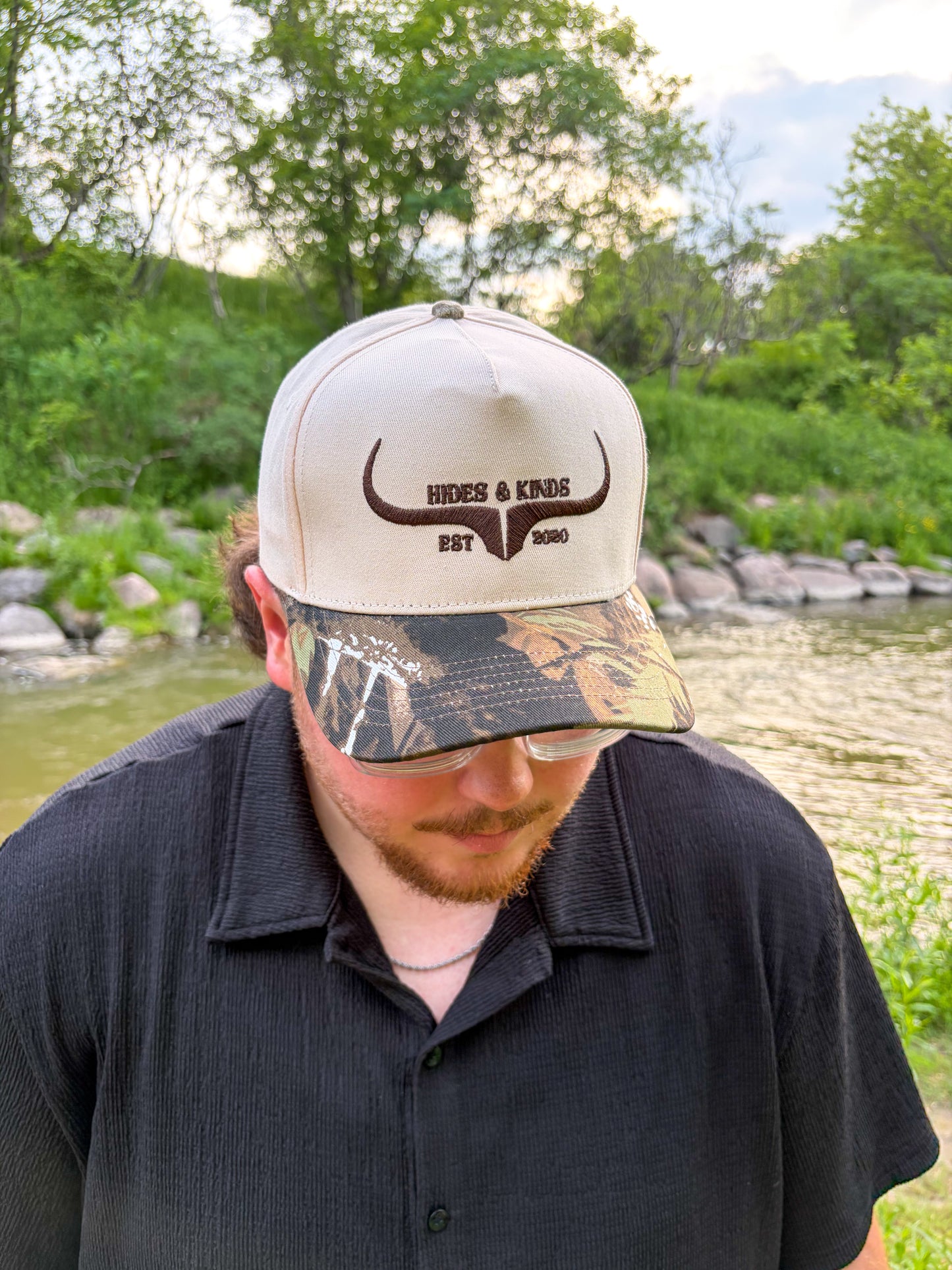 Man wearing the Hides & Kinds Original Logo Hat in camo with a cream crown and brown embroidered logo.
