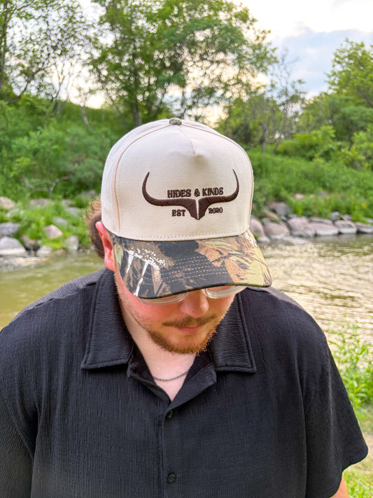 Man wearing the Hides & Kinds Original Logo Hat in camo with a cream crown and brown embroidered logo.