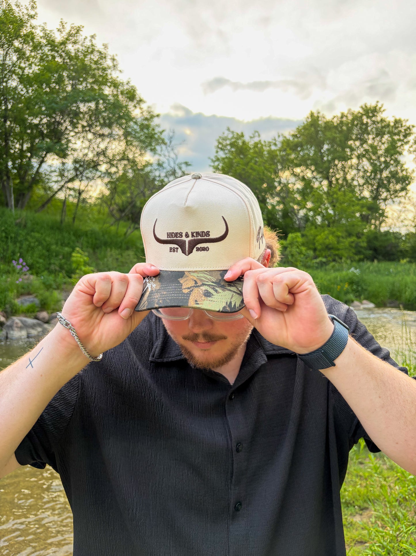 Lifestyle photo of the Hides & Kinds Original Logo Hat (Camo) featuring a cream crown, camo brim, and brown embroidered logo.