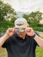Lifestyle photo of the Hides & Kinds Original Logo Hat (Camo) featuring a cream crown, camo brim, and brown embroidered logo.