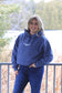 Full front facing view of a woman wearing the H&K Original Logo Hoodie in denim blue with natural forest scenery in the background.
