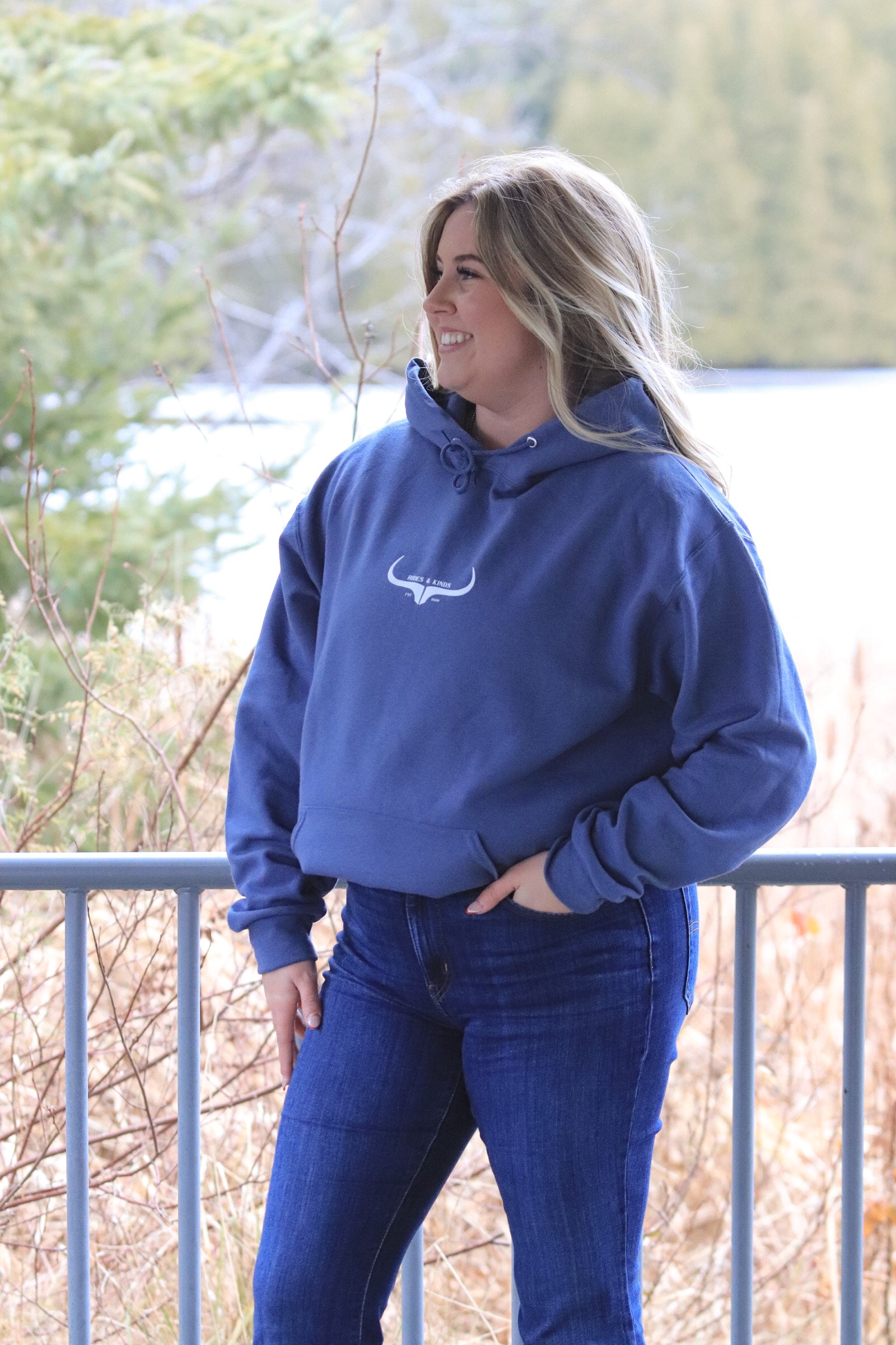Woman looking to the side, standing outdoors in the Original Logo Hoodie (denim blue), showing the relaxed, cozy fit.