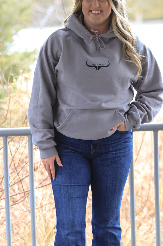 Close-up of grey Hides and Kinds hoodie featuring bullhorn logo, model smiling with one hand in the front pocket.