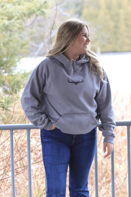 Woman wearing grey Hides and Kinds hoodie looking to the right, standing outdoors by a railing with trees in the background.
