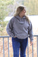 Woman wearing grey Hides and Kinds hoodie looking to the right, standing outdoors by a railing with trees in the background.