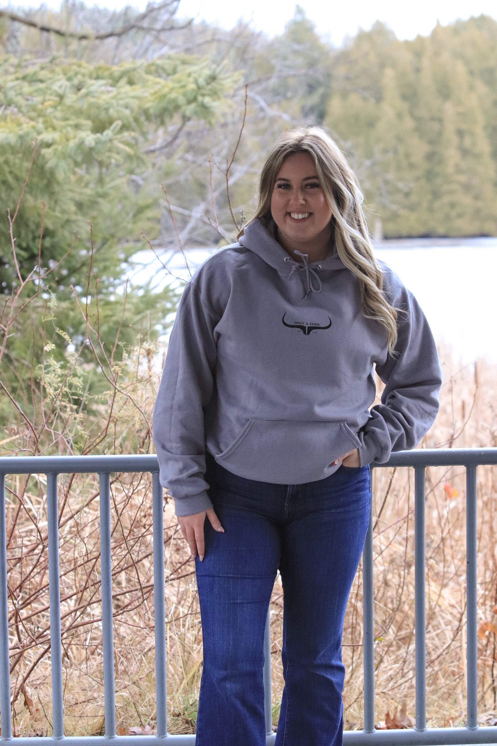 Full body shot of woman in grey Hides and Kinds hoodie and blue jeans, posing in front of a forest and lake backdrop.