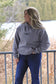 Model smiling and looking to the side in a grey Hides and Kinds hoodie, surrounded by natural outdoor scenery.