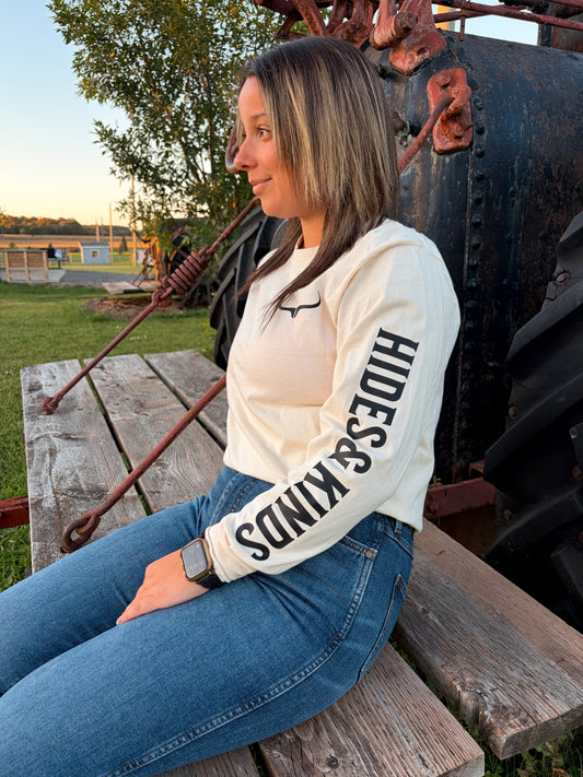 Model wearing the cream Hides & Kinds Original Logo Long Sleeve with black lettering down the sleeve and a small longhorn logo on the chest, seated outdoors.