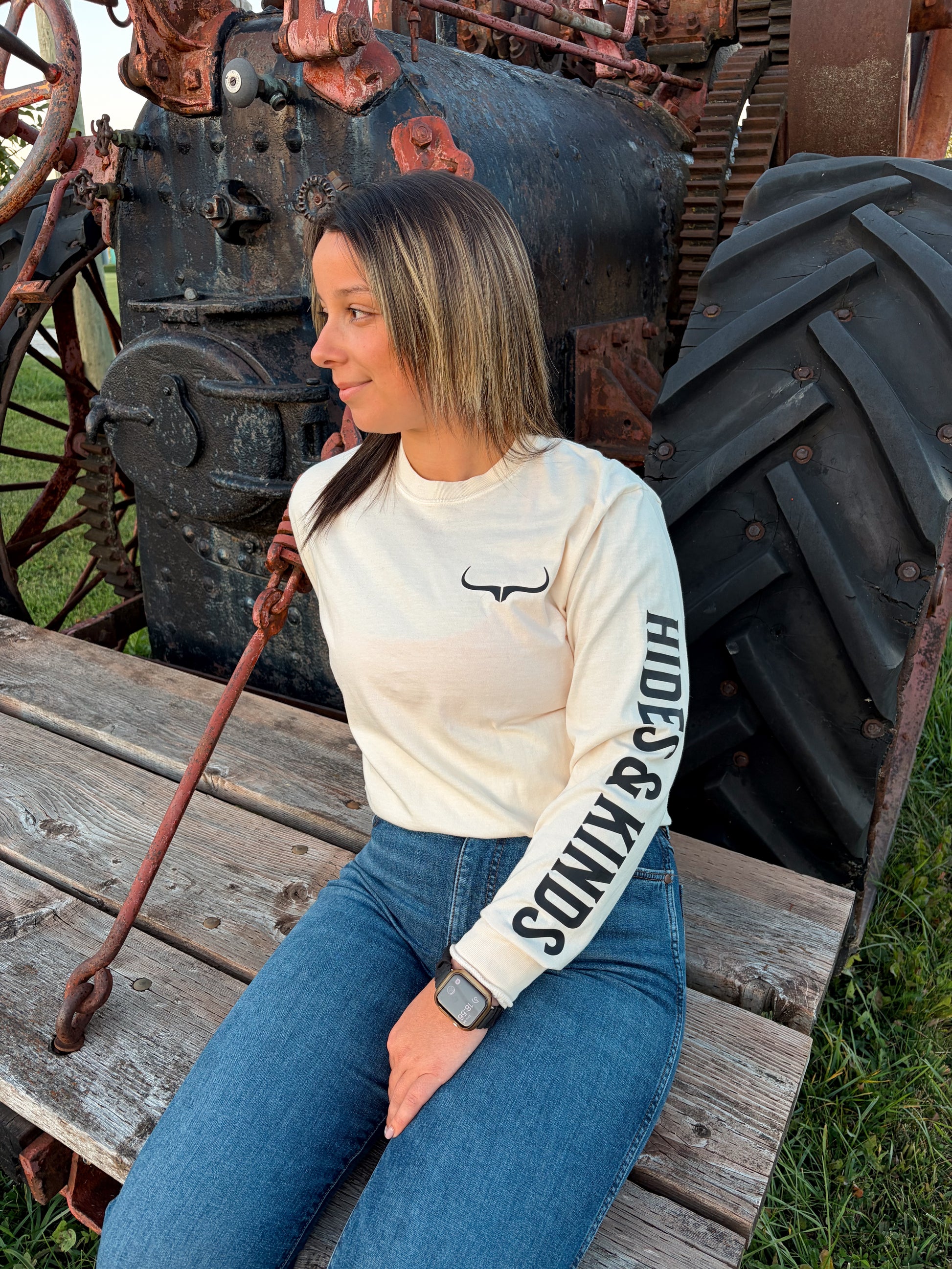 Front view of the cream Original Logo Long Sleeve by Hides & Kinds, showing the black longhorn chest logo and bold Hides & Kinds sleeve print, styled with blue jeans.