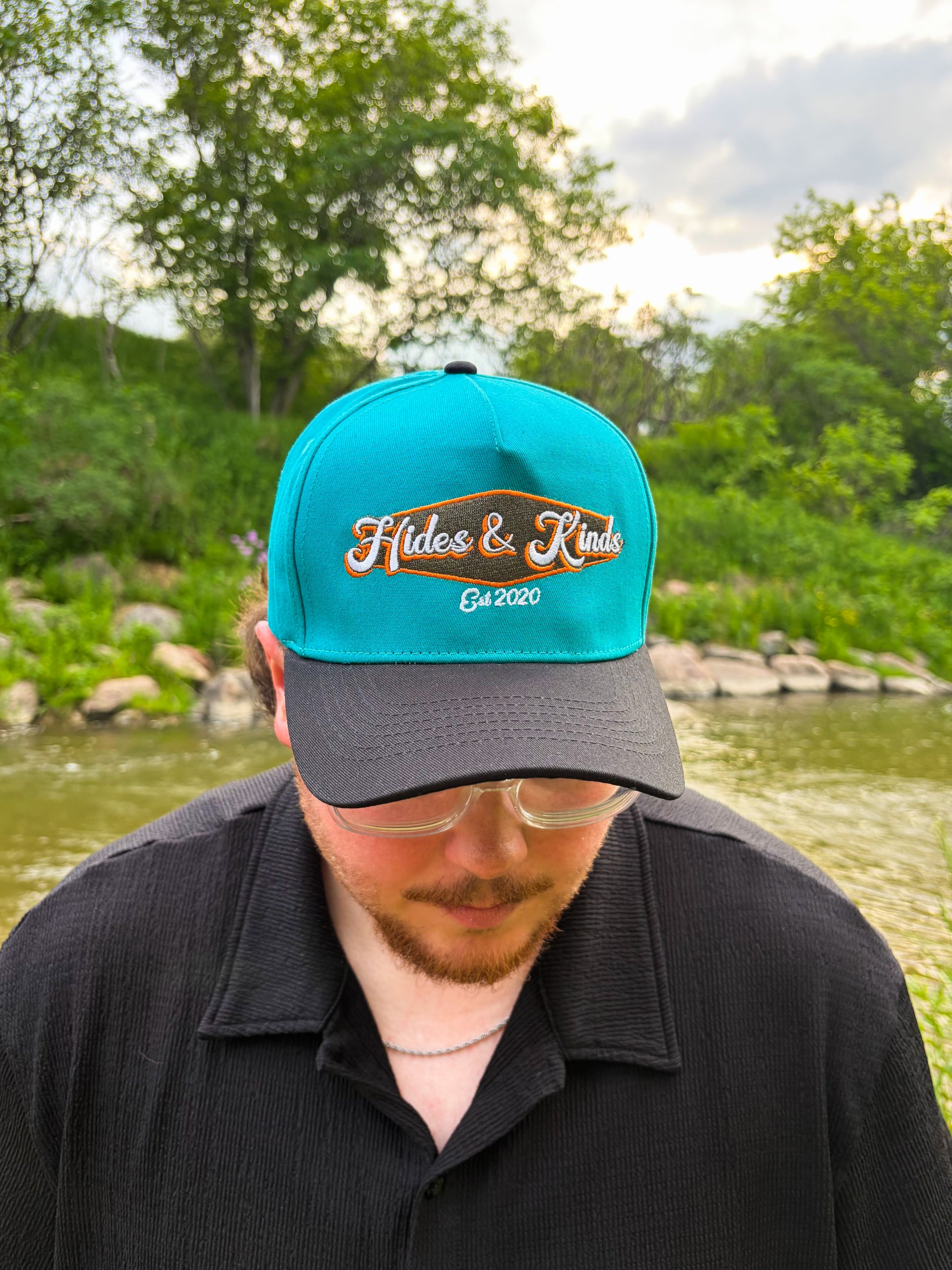 Man wearing the Hides & Kinds Retro Country Hat with a teal crown, black brim, and vintage inspired embroidered logo.