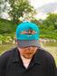 Man wearing the Hides & Kinds Retro Country Hat with a teal crown, black brim, and vintage inspired embroidered logo.