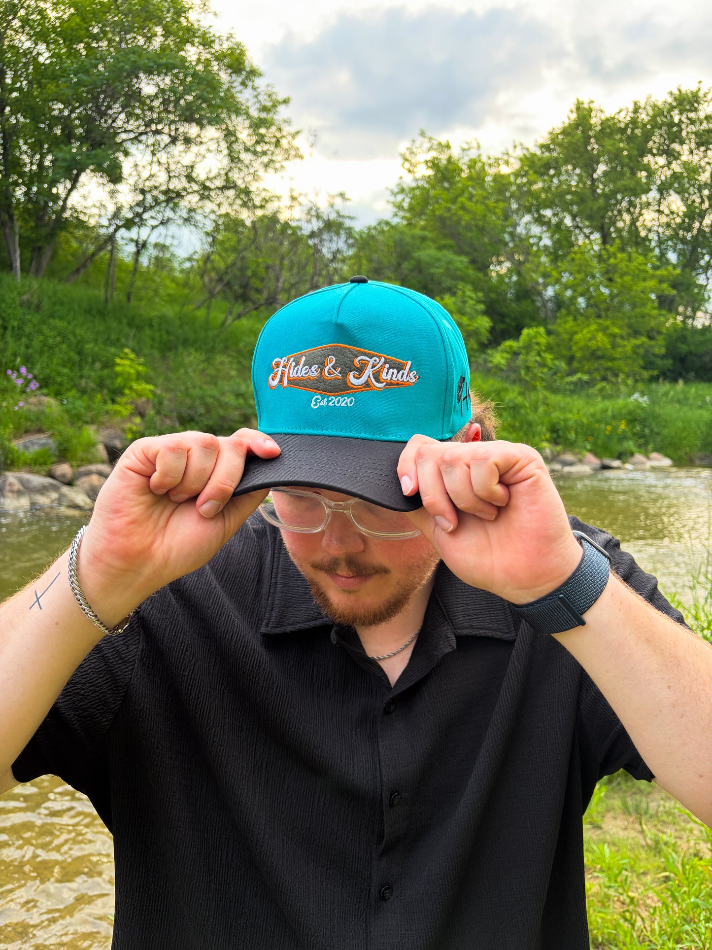 Lifestyle photo of the Hides & Kinds Retro Country Hat featuring a teal crown, black brim, and embroidered Hides & Kinds logo.