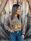 Model wearing the brown Hides & Kinds Stockman Crewneck with blue jeans and a western belt, standing in front of a rustic wooden barn.
