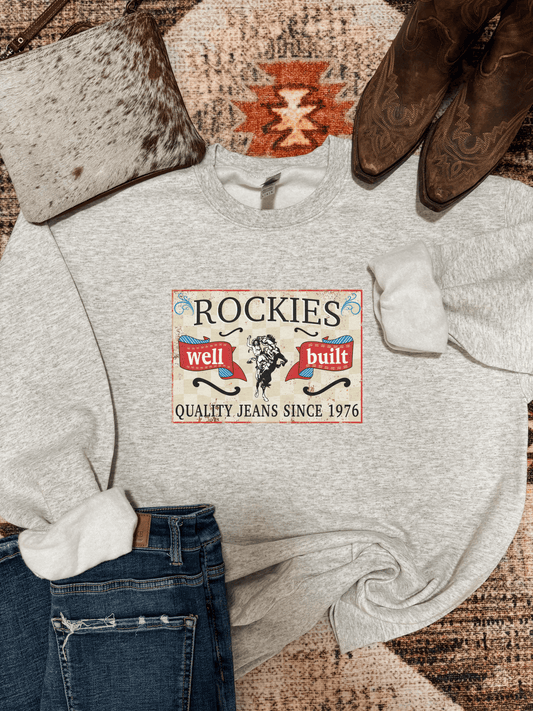 Flat lay of the Vintage Rockies Crewneck styled with distressed denim, cowhide purse, and cowboy boots on a southwestern rug