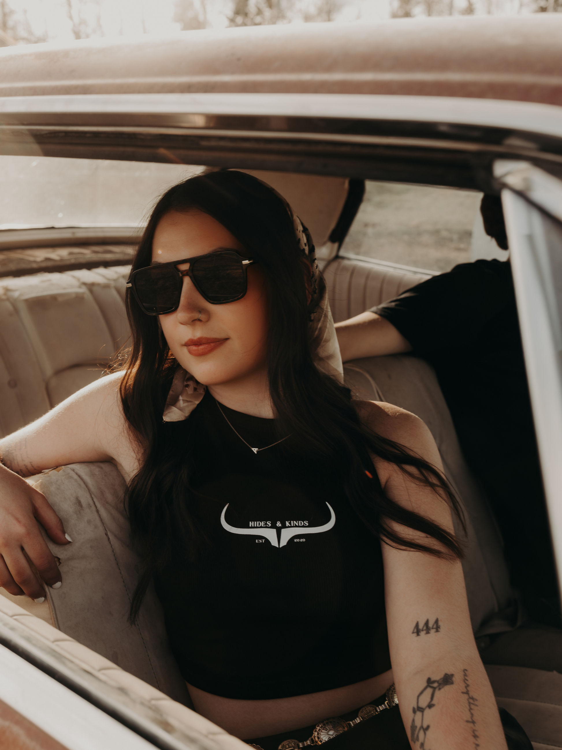 Woman sitting in a vintage car wearing a black Hides & Kinds cropped high neck ribbed tank with original western logo.
