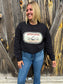 Full body shot of smiling model wearing Western Harvest crewneck by Hides & Kinds, standing in front of wood wall.