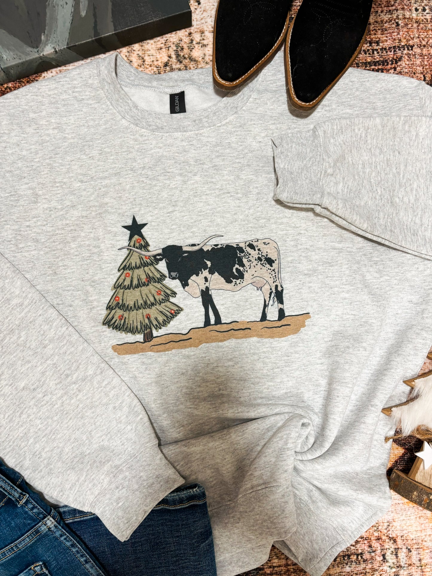 Close up of a Christmas themed longhorn graphic crewneck, flat lay with black boots and canvas artwork – cozy western holiday look.